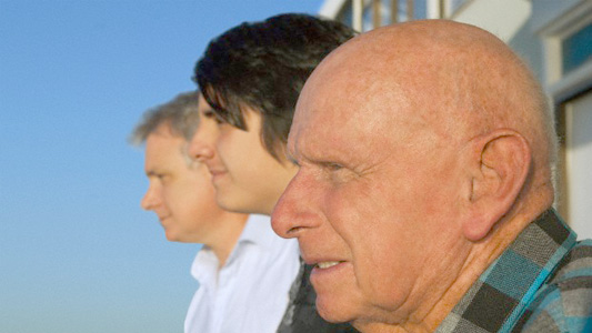 Na foto, o perfil do rosto de três homens, de idades diferentes, com um céu azul de fundo